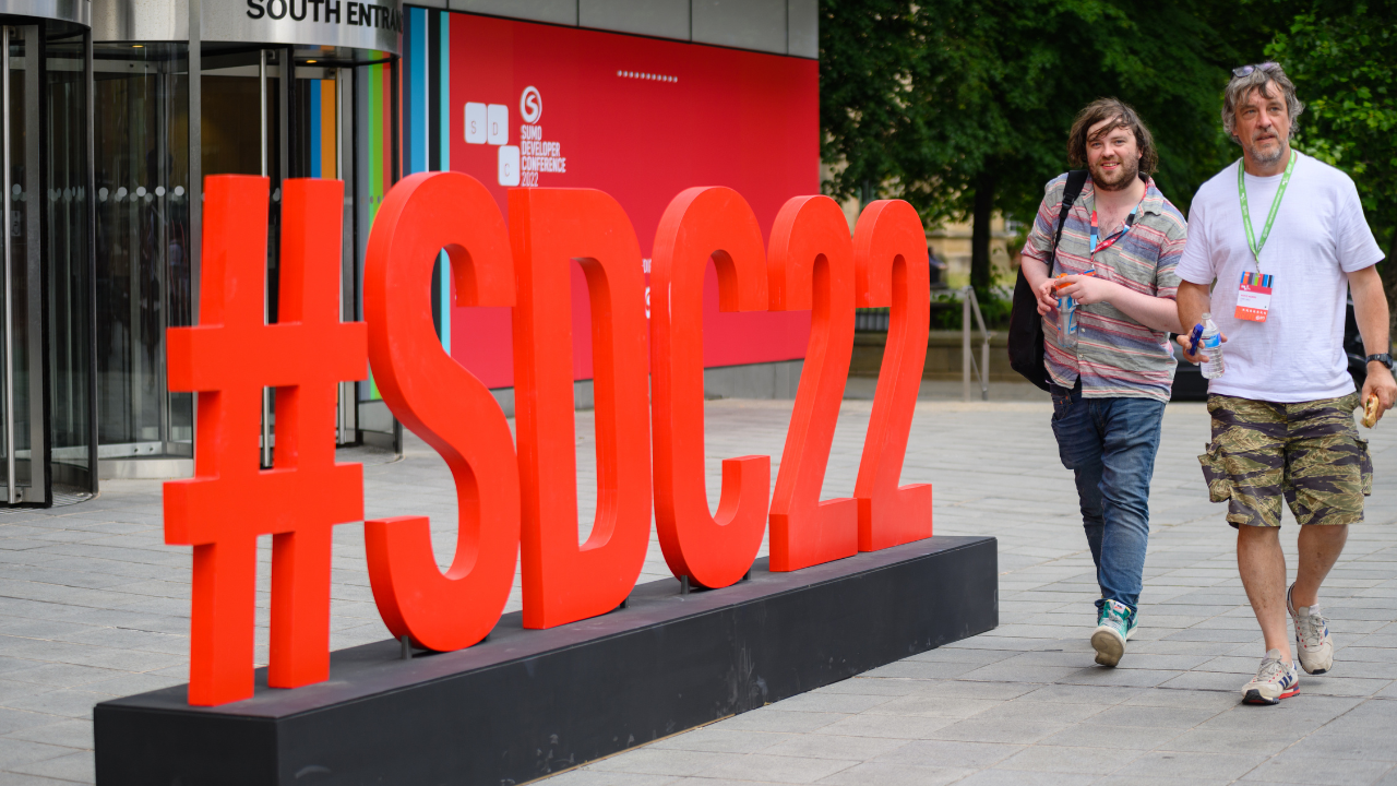 Two men walking past a large freestanding sign which reads #SDC22